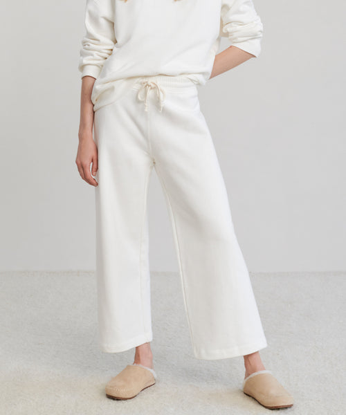 Wearing the Marina Sweatpant—loose-fitting, wide-leg cotton loungewear—with a matching white sweatshirt and beige slippers, this person stands on a light carpeted floor against a plain background. Their face is not visible.