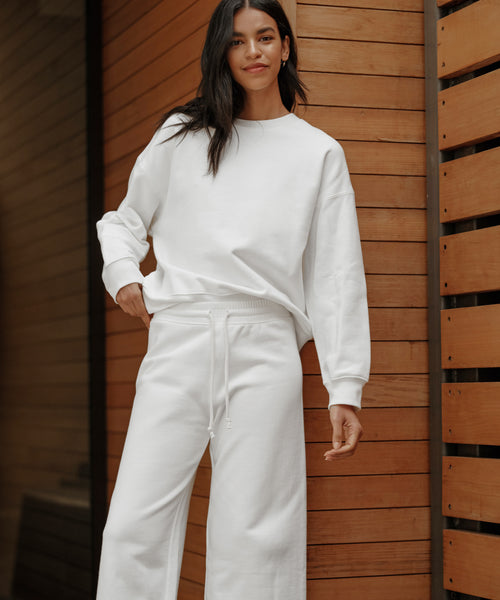 A woman stands outdoors by a wooden wall, smiling slightly with her hand in her pocket, wearing relaxed cotton loungewear—a white sweatshirt and the Marina Sweatpant in a matching wide-leg style.