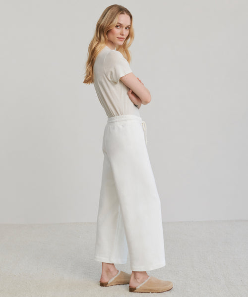 A woman with long blonde hair stands sideways, facing the camera. She wears a beige short-sleeve top, tan slip-on shoes, and white Marina Sweatpants. The background is plain and light-colored.