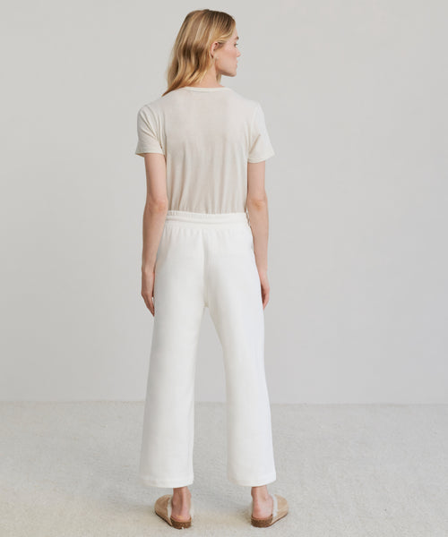 A woman with blonde hair, seen from behind, wears a short-sleeve beige cotton t-shirt, Marina Sweatpant in white with an elastic waistband, and slippers against a plain, light background.