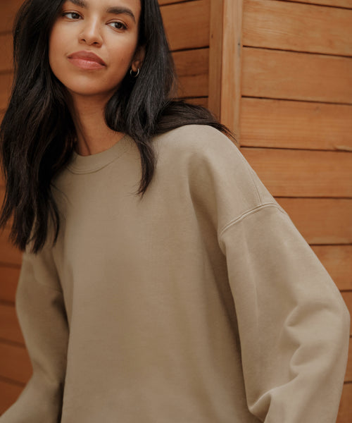 A woman with long dark hair wears the Marina Sweatshirt and stands before a wooden wall, gazing slightly to the side with a soft, relaxed expression.