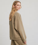 A woman with long blonde hair models the 100% cotton, oversized, light brown Marina Sweatshirt and matching pants, standing sideways and looking over her shoulder against a plain light background.