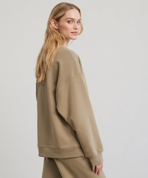 A woman with long blonde hair models the 100% cotton, oversized, light brown Marina Sweatshirt and matching pants, standing sideways and looking over her shoulder against a plain light background.