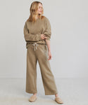 A woman stands in a neutral-toned room wearing the relaxed Marina Sweatshirt, crafted from 100% cotton—perfect for lounging. She pairs it with wide-leg sweatpants and beige slippers, looking to the side with effortless calm.