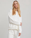 A woman with long blonde hair stands confidently against a plain light background, wearing the loose-fitting Marina Sweatshirt—crafted from 100% cotton—and matching white pants.