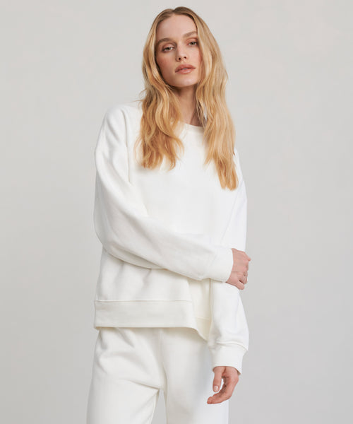 A woman with long blonde hair stands confidently against a plain light background, wearing the loose-fitting Marina Sweatshirt—crafted from 100% cotton—and matching white pants.