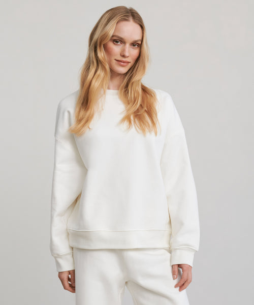 A person with long, wavy blonde hair wears the loose-fitting Marina Sweatshirt—crafted from 100% cotton—with matching white pants, standing against a plain light background and smiling softly at the camera.