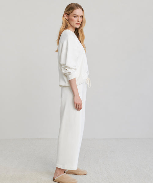 A woman with long blonde hair stands sideways, barefoot on a light carpet, wearing the Marina Sweatshirt and matching wide-leg pants—an effortless 100% cotton loungewear set. The background is plain white.