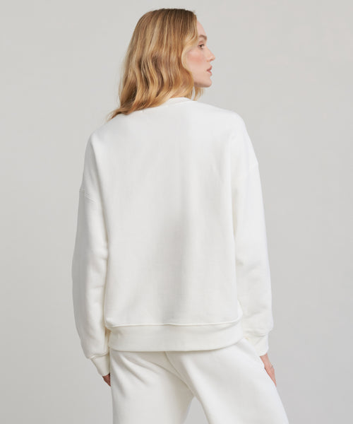 A woman with long, wavy blonde hair is seen from behind, wearing the loose-fitting, 100% cotton Marina Sweatshirt and matching white sweatpants against a plain light background.