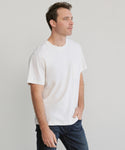 A man with short brown hair wears the Mens Vintage Pocket Tee in white and blue jeans, standing with one hand in his pocket and looking off to the side against a neutral background.