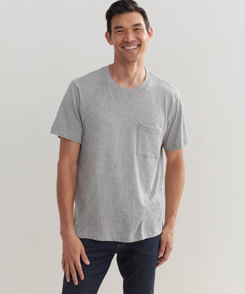 A smiling man stands against a plain light background, wearing the Mens Vintage Pocket Tee in grey with a chest pocket, paired with dark jeans.