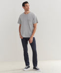 A man stands against a plain light wall, wearing the Mens Vintage Pocket Tee in grey, paired with dark jeans and black high-top sneakers. He looks slightly to the side with a relaxed expression.