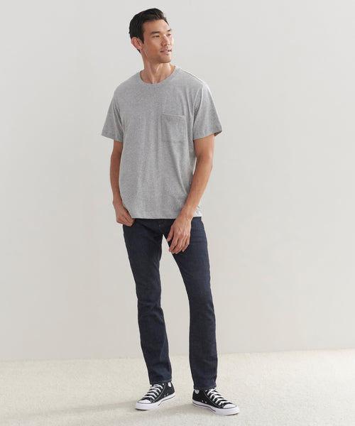A man stands against a plain light wall, wearing the Mens Vintage Pocket Tee in grey, paired with dark jeans and black high-top sneakers. He looks slightly to the side with a relaxed expression.