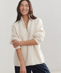 A woman with long dark hair smiles, standing with one arm crossed. She wears the Oversized Half Zip in ivory and dark pants against a plain light background.