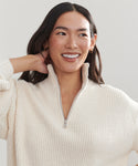 Woman with long dark hair wears the Oversized Half Zip in ivory boucle rib, smiling and looking slightly to the side with one hand behind her head against a neutral background.