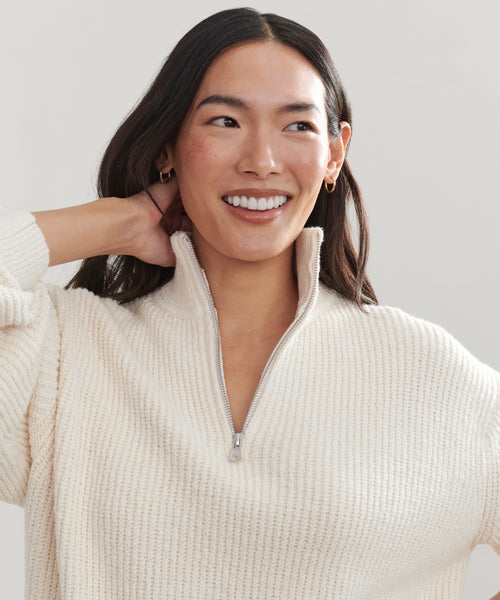Woman with long dark hair wears the Oversized Half Zip in ivory boucle rib, smiling and looking slightly to the side with one hand behind her head against a neutral background.