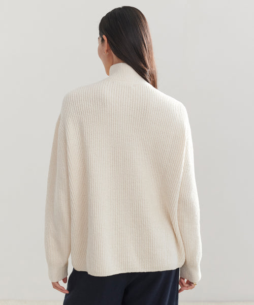 A person with long dark hair, seen from behind, wears an ivory Oversized Half Zip sweater and dark pants, standing before a plain light background.