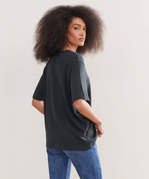 A woman with voluminous curly hair models the Relaxed Tee in black with blue jeans, standing sideways and looking over her shoulder against a plain light background.