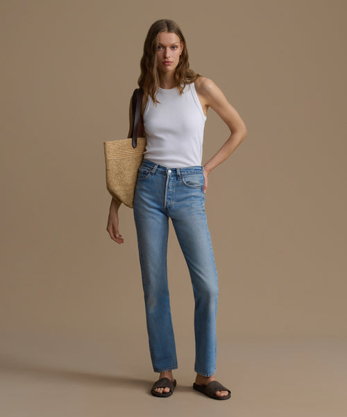 Woman wearing a white tank top and blue jeans, holding a woven bag against a beige background