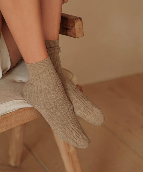 Person sitting on a wooden chair with legs crossed, wearing Rib Socks; the cozy setting and warm lighting highlight these all-season socks, ideal for everyday comfort on neutral-toned floors.