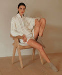 A woman with dark hair sits calmly on a wooden chair, wearing Rib Socks alongside a chunky white cable-knit sweater and white shorts, resting one arm and foot on the chair as she looks at the camera.