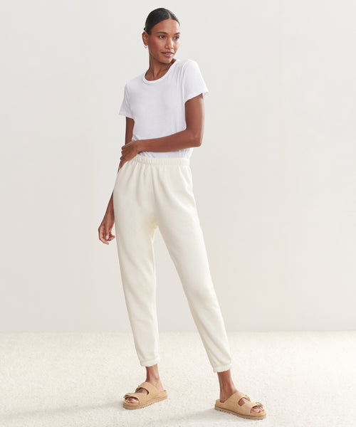 A woman stands in a minimal, sunlit room wearing a soft white tee and the Saturday Sweatpant joggers with tan sandals. Her hair is pulled back and she gazes to the side with a relaxed expression.