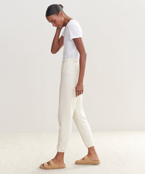 A woman stands sideways, looking down with one hand on her neck. She wears an ultra-soft white tee, off-white Saturday Sweatpant joggers, and beige sandals against a plain light background.
