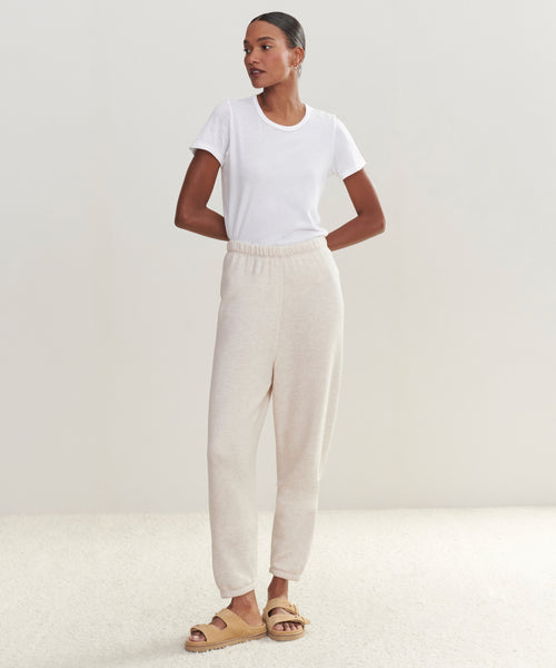 Against a plain background, a woman with tied-back hair and crossed arms looks to the side, wearing light beige Saturday Sweatpant joggers, an ultra-soft white tee, and tan sandals.