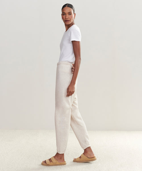 A woman stands sideways wearing a white t-shirt, light beige Saturday Sweatpant joggers, and tan slide sandals against a plain, light-colored background.