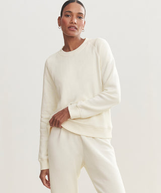 Someone models the cream Saturday Sweatshirt, made of soft French terry, paired with matching sweatpants. They stand against a plain, light background and look confidently at the camera, one hand on their hip.