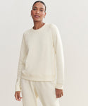 A woman in a cream Saturday Sweatshirt, made from soft French terry, stands against a plain light background and looks at the camera with a neutral expression.