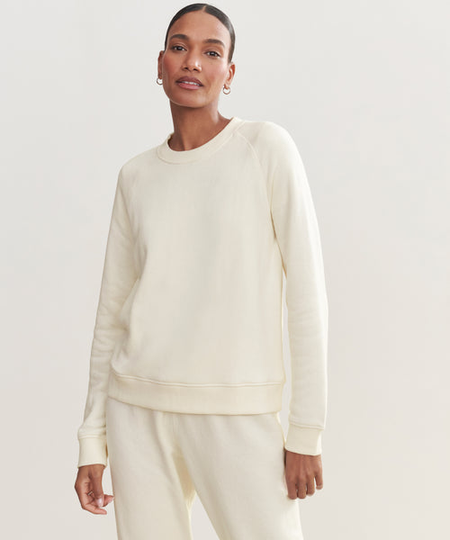 A woman in a cream Saturday Sweatshirt, made from soft French terry, stands against a plain light background and looks at the camera with a neutral expression.