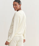 A woman in a cream Saturday Sweatshirt and matching French terry pants stands turned slightly to the side, smiling over her shoulder. The background is plain and light-colored.