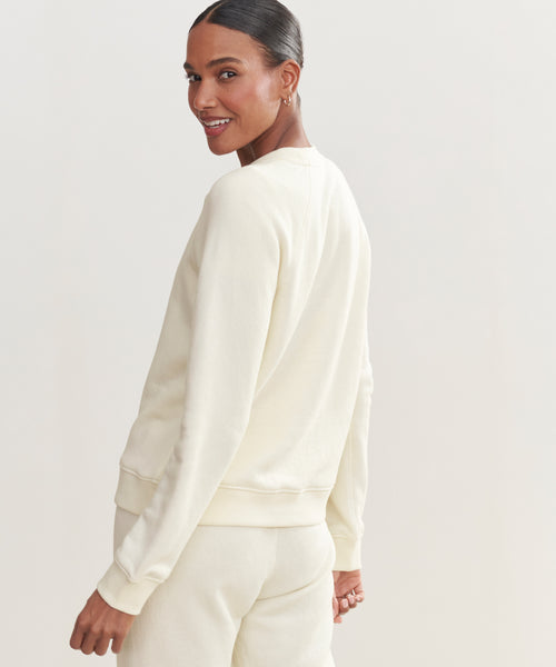 A woman in a cream Saturday Sweatshirt and matching French terry pants stands turned slightly to the side, smiling over her shoulder. The background is plain and light-colored.