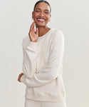 A woman smiles warmly, touching her face and wearing the light beige Saturday Sweatshirt in soft French terry with matching pants, set against a simple, light background.