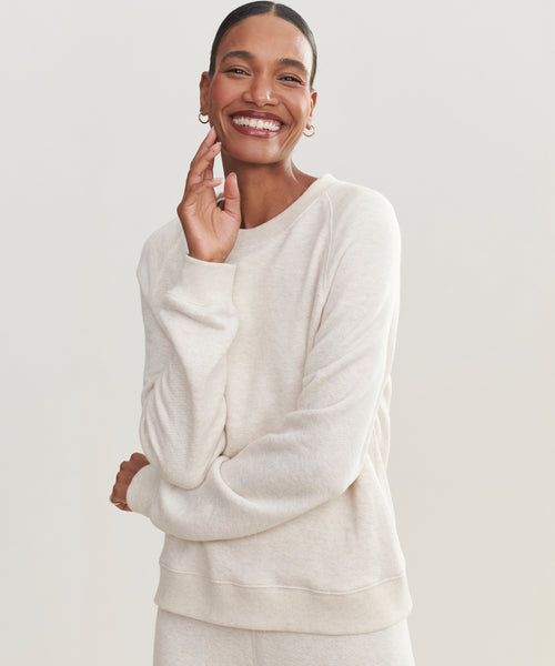 A woman smiles warmly, touching her face and wearing the light beige Saturday Sweatshirt in soft French terry with matching pants, set against a simple, light background.