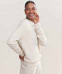 A woman smiles in a light beige Saturday Sweatshirt made from soft French terry, paired with matching pants. She stands against a plain light background, one hand on her hip and the other near her face.