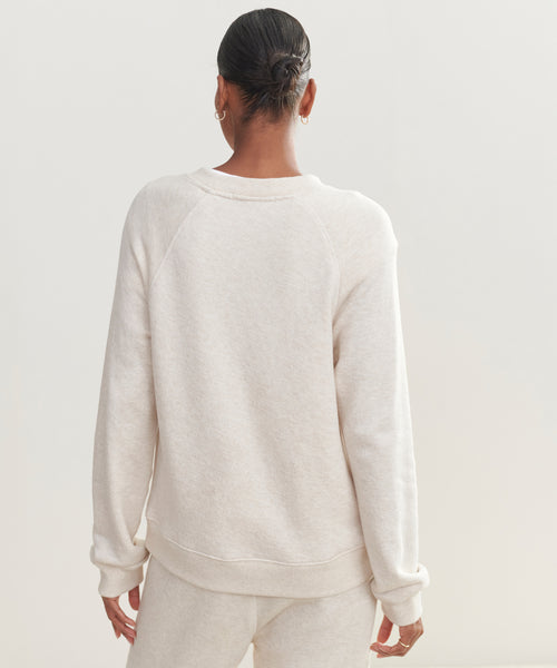 A person with dark hair in a bun is seen from behind, wearing the light beige Saturday Sweatshirt and matching soft French terry pants, standing against a plain light background.