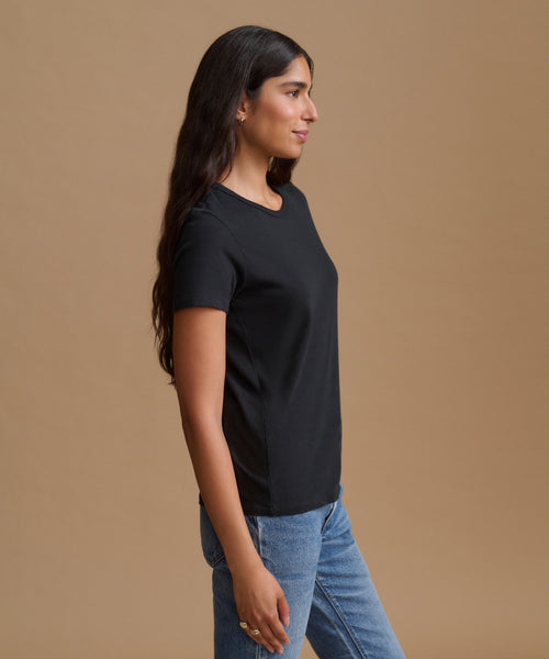 A woman in a black short-sleeve crewneck t-shirt and blue jeans, shown in profile against a solid tan studio background.