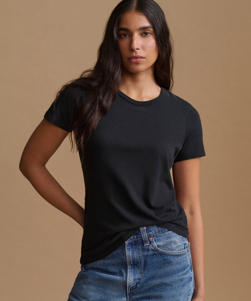 A woman modeling a black short-sleeve crewneck t-shirt paired with blue jeans, set against a solid warm beige studio background.