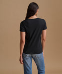 A woman viewed from the back wearing a plain black short-sleeve crew-neck T-shirt and blue jeans, standing against a solid warm tan studio background.