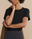 A woman in a fitted black Cotton Slim Tee and a dark pleated skirt stands with one hand on her chin, draping a tan coat over her shoulder against a beige background. Her face is partially out of frame.