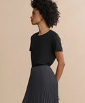 A woman with curly hair stands in profile against a beige background, wearing the Cotton Slim Tee—a black, crewneck, fitted top—paired with a high-waisted, pleated dark skirt.