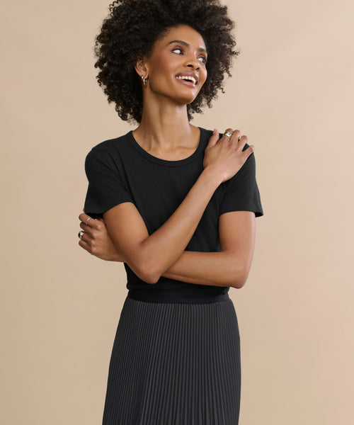 A woman with curly hair wears the Cotton Slim Tee in black and a black pleated skirt, standing against a beige background, smiling and looking to the side with her arms gently crossed.