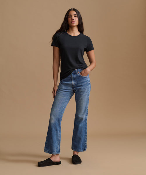A woman modeling a black short-sleeve t-shirt and blue high-waisted straight-leg jeans against a solid beige studio background.