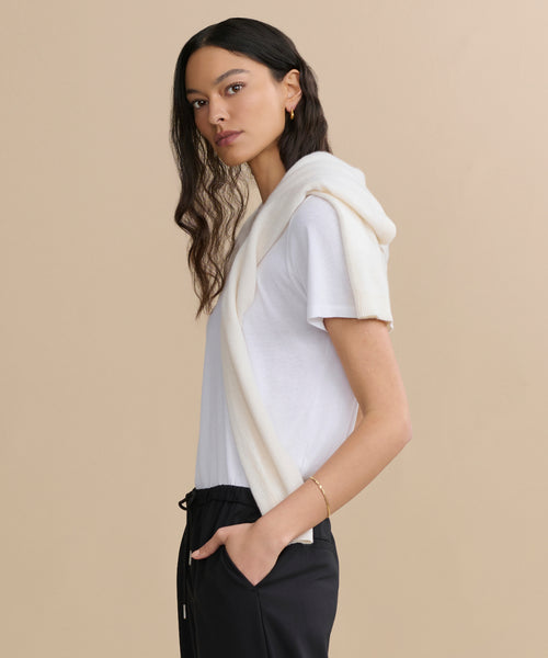 A woman with long, wavy dark hair stands sideways, wearing the Cotton Slim Tee in white, paired with black pants and a cream sweater over her shoulders. She has a neutral expression and stands against a minimal background.