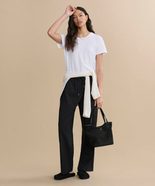 A woman stands against a beige background wearing the Cotton Slim Tee in white, black pants, and shoes, with a cream sweater tied at her waist. She holds a black handbag and touches her head with one hand.