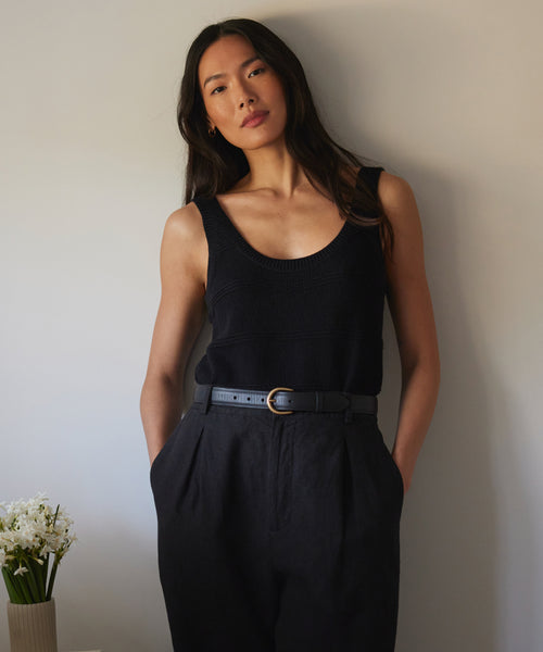 A woman with long dark hair stands by a neutral wall, wearing a Sweater Tank tucked into high-waisted black pants with a belt. A table to her left holds a small vase of white flowers.