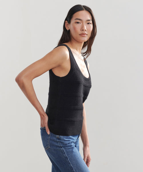 A woman with long dark hair wears a Sweater Tank in black and blue jeans, standing against a plain light background, facing slightly to the side with a neutral expression.