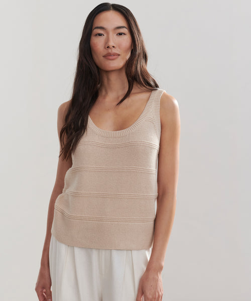 A woman with long dark hair wears the Sweater Tank, made from 100% cotton, paired with loose white pants and stands against a plain light background.